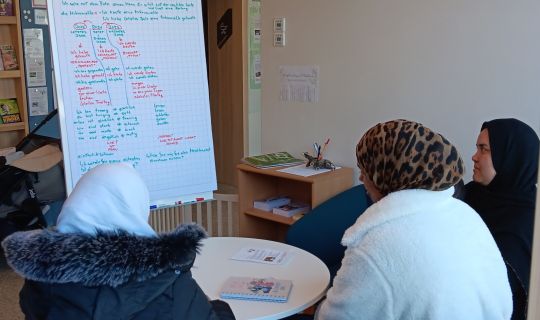 Drei Frauen sitzen am Tisch und schauen auf ein Flipchart. Drei Frauen sitzen am Tisch und schauen auf ein Flipchart.