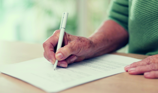 Hand einer älterwerdenden Person schreibt mit einem Stift auf einem Blatt Papier Hand einer älterwerdenden Person schreibt mit einem Stift auf einem Blatt Papier